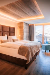 Premium Chic Room mit Doppelbett, türkisem Sessel, Holzdecke und Balkonblick auf Chalets und Berge, Hotel Bellerive Zermatt.