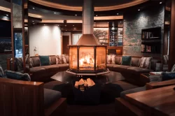 Lounge mit rundem Sofa, türkisen Kissen und gläsernem Kaminfeuer im Hotel Bellerive Zermatt