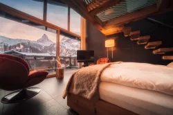 Gemütlicher Sky Room: weisses Bett, roter Sessel und Holztreppe, Matterhorn und Zermatt-Lichter durch die Fenster, Hotel Bellerive Zermatt.