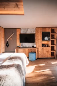 Helles Deluxe-Zimmer mit Holzwänden, Bett, TV-Wand und Balkonblick auf Schnee und Chalets im Hotel Bellerive Zermatt.