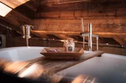 Freistehende Badewanne mit Holztablett, Badesalz und Herzseife vor Holzwänden im Sonnenlicht, Hotel Bellerive Zermatt.