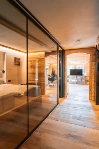 Holzkorridor mit Glaswand zum Bett und Blick ins Wohnzimmer im Apartment The Alpine Nest 1895, Hotel Bellerive Zermatt.